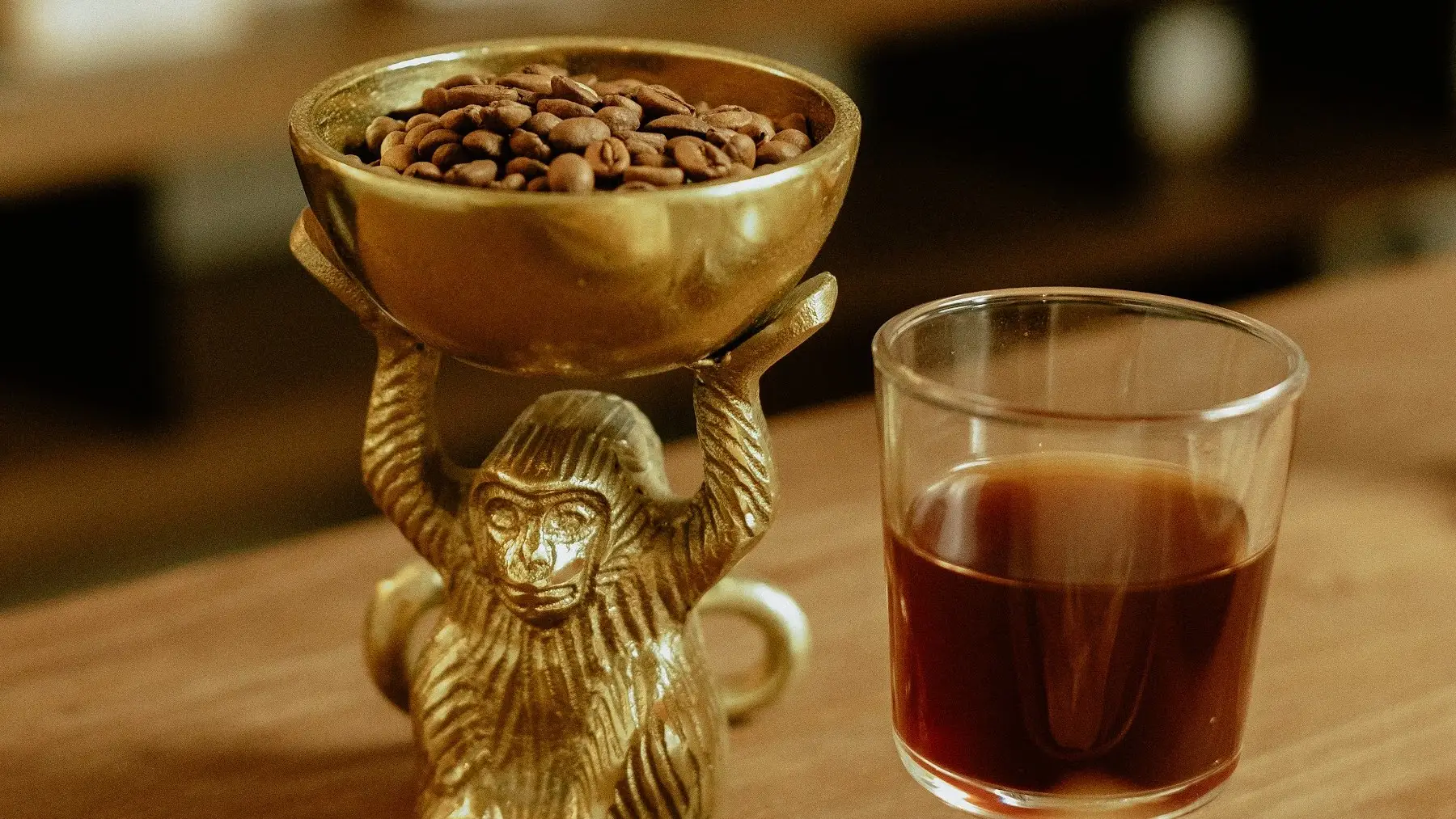 Un mono dorado sostiene un cuenco de granos de café junto a un vaso de café de filtro